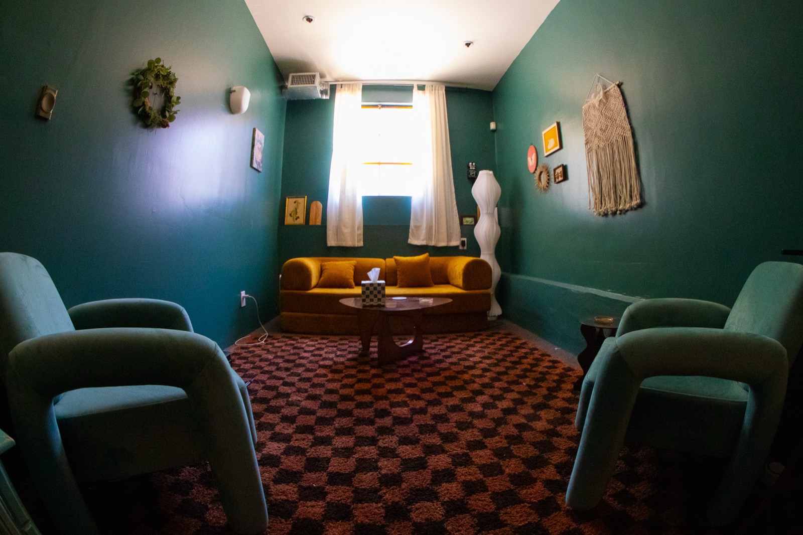 A therapy room with deep green walls, a mustard yellow velvet sofa under a curtained window, two pale green armchairs facing it, a small round table with a box of tissues, and a brown checkerboard rug.